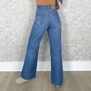 Può includere: Jeans blu in denim con gamba svasata. I jeans hanno una vita alta e un classico design a cinque tasche. Una piccola etichetta arancione e bianca è visibile sulla tasca posteriore. I jeans sono di lavaggio medio e sembrano vintage.