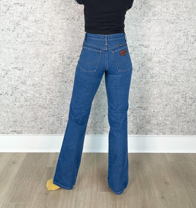 May include: Blue denim Wrangler jeans with a flared leg. The jeans have a high waist and a brown leather patch on the back pocket. The person is wearing a black top. The jeans are a classic style.
