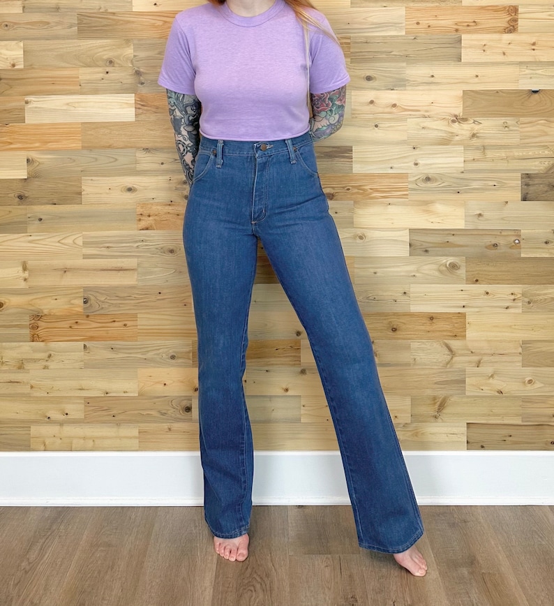 Pu&ograve; includere: Una maglietta girocollo lilla chiaro e jeans di denim blu scuro a vita alta. I jeans hanno una gamba svasata. L'outfit &egrave; esposto contro un muro di assi di legno e un pavimento marrone chiaro.