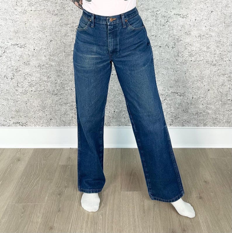 May include: Dark wash denim jeans with a high waist and a wide leg. The jeans have a classic five-pocket design and a button and zipper closure. The model is wearing white socks.