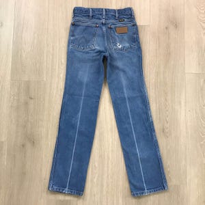 May include: A pair of blue denim jeans featuring a classic five-pocket design, a high-rise waist, and a straight-leg cut. The back pockets display a signature stitched W pattern, a brown leather brand patch, and a small distressed hole on the right.