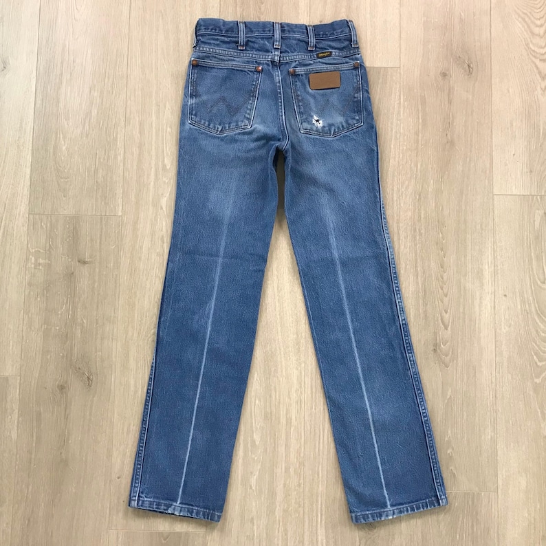 May include: A pair of blue denim jeans featuring a classic five-pocket design, a high-rise waist, and a straight-leg cut. The back pockets display a signature stitched W pattern, a brown leather brand patch, and a small distressed hole on the right.