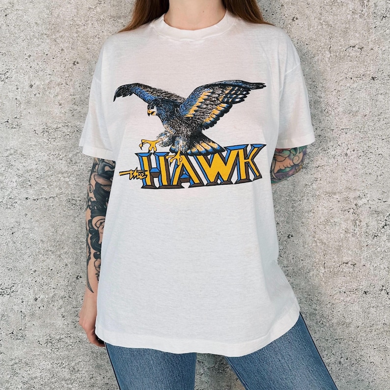 May include: White t-shirt featuring a detailed graphic of a hawk with outstretched wings. The hawk is depicted in shades of blue, yellow, and gray, perched above the text "the HAWK" in a bold, stylized font. A vintage-style tee.