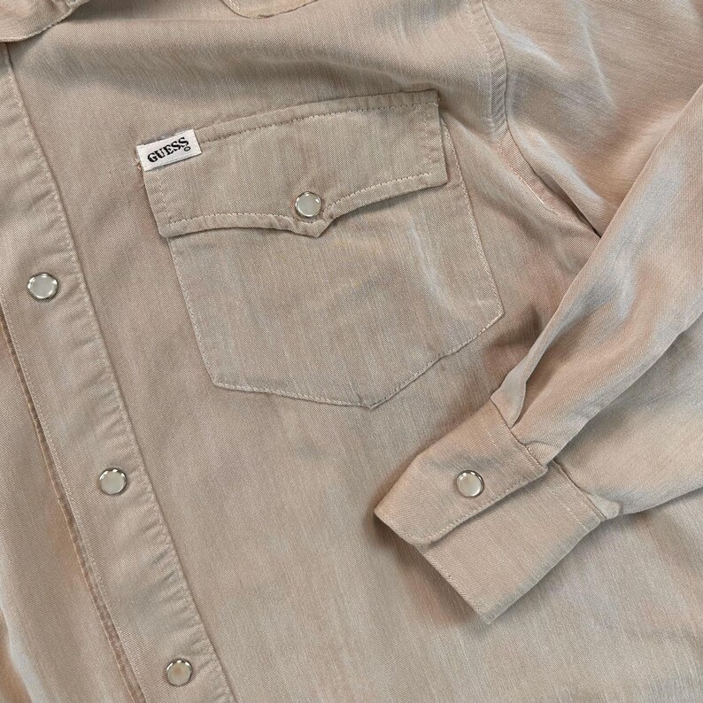 May include: A tan-colored button-up shirt with a pocket on the chest. The shirt has a Guess label and silver-colored buttons. The long sleeves have buttoned cuffs. The fabric has a subtle texture.