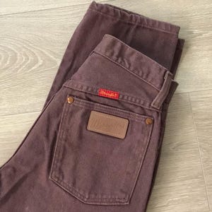 Op de afbeelding: Een bruine Wrangler-jeans. De jeans heeft een bruin leren label met het Wrangler-logo op de achterzak en een rood label met het Wrangler-logo. De jeans is opgevouwen op een lichtbruin houten oppervlak.