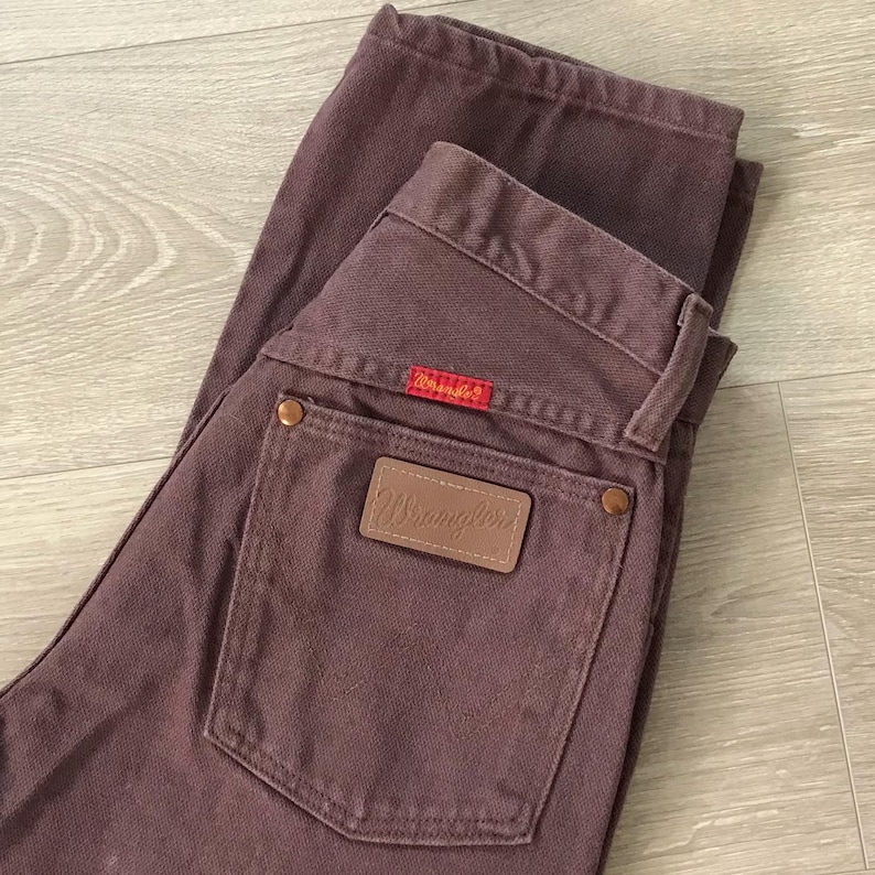 Op de afbeelding: Een bruine Wrangler-jeans. De jeans heeft een bruin leren label met het Wrangler-logo op de achterzak en een rood label met het Wrangler-logo. De jeans is opgevouwen op een lichtbruin houten oppervlak.