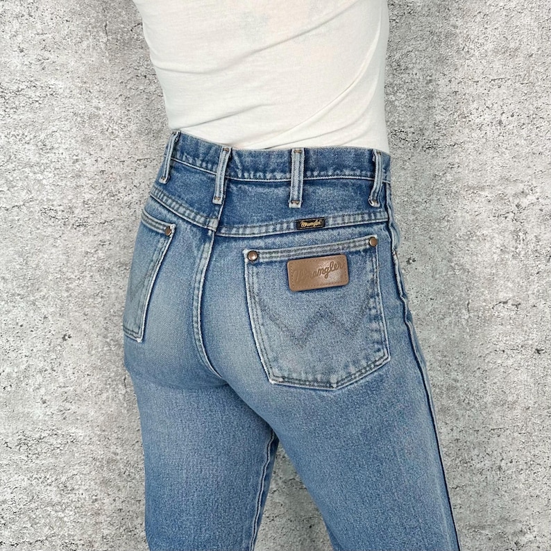 May include: Light blue denim jeans with a brown leather Wrangler patch on the back pocket. The jeans have a classic five-pocket design and a high waist. The jeans are worn with a white top.