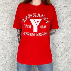 May include: A red t-shirt with white text and a graphic. The text reads "KANKAKEE" above a white "Y" logo with "YM" and "CA" on either side. Below the logo, the text reads "SWIM TEAM".