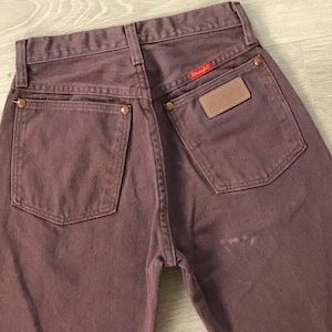 Op de afbeelding: Close-up van de achterkant van een stofroze Wrangler-jeans. De jeans heeft twee achterzakken, koperen klinknagels en een rood Wrangler-label. Een bruine leren patch met het Wrangler-logo is zichtbaar.