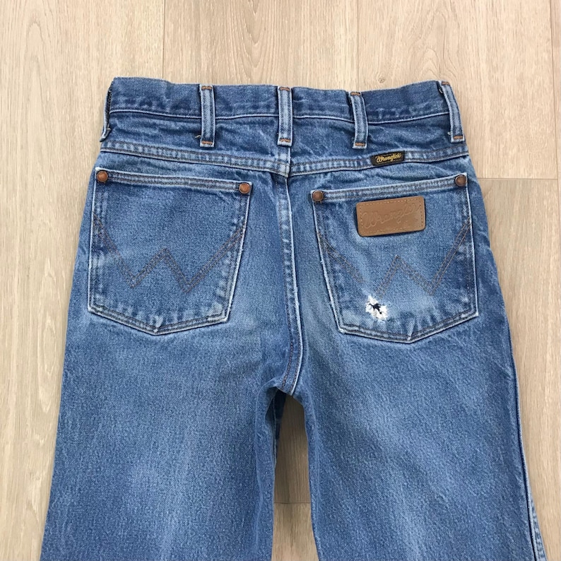 May include: Blue denim jeans featuring a high-rise waist, belt loops, and two back pockets with a signature W-shaped stitch pattern. A brown leather Wrangler brand patch is attached to the right pocket, which also displays a small frayed hole.