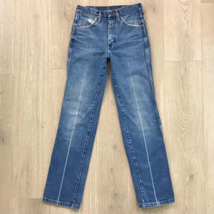 May include: A pair of high-waisted blue denim jeans featuring a classic five-pocket design, a button fly closure, and a straight-leg cut. The fabric displays a faded wash with subtle whiskering details along the thighs and front pockets.
