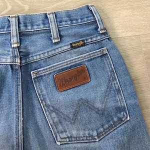 Op de afbeelding: Close-up van lichtblauwe denim jeans met een bruine leren Wrangler patch op de achterzak. De jeans heeft klassieke stikdetails en een Wrangler label boven de achterzak.