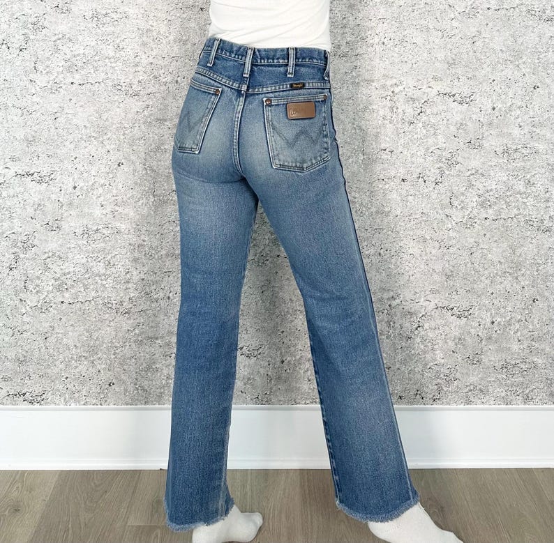 May include: Blue denim jeans with a classic fit, featuring a high waist and a straight leg. The jeans have a brown leather patch on the back pocket and the Wrangler logo. The jeans are worn and have a faded look.