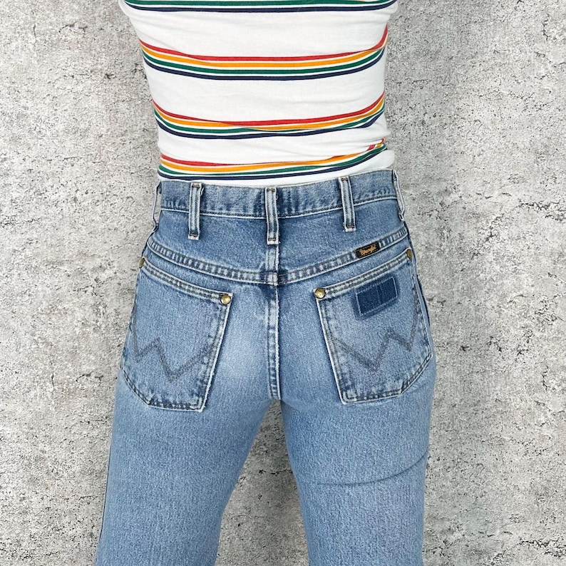 Puede incluir: Vaqueros Wrangler de mezclilla azul claro con cintura alta y dise&ntilde;o cl&aacute;sico de bolsillo trasero. Los vaqueros presentan el logotipo de Wrangler en el bolsillo trasero y se combinan con una camiseta a rayas con colores del arco&iacute;ris.