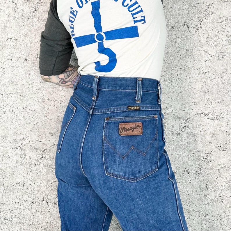 May include: Blue denim Wrangler jeans with a brown leather patch on the back pocket. The jeans have a high waist and a classic fit. The person is wearing a white t-shirt with blue text that says "BLUE OF CULT" and gray sleeves.