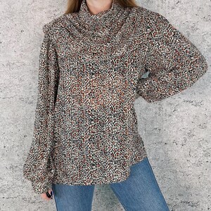 May include: A long-sleeved blouse with a cowl neck and a leopard print pattern in shades of brown, black, and cream. The blouse has a relaxed fit and is paired with blue jeans.