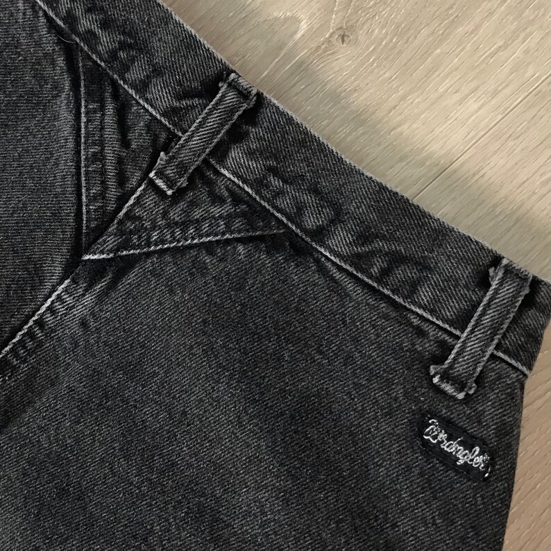 Pu&ograve; includere: Primo piano di una sezione di jeans neri. I jeans hanno un design classico con cuciture visibili e passanti per cintura. Il logo Wrangler &egrave; visibile su una piccola toppa nera.
