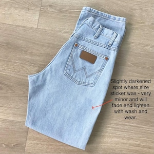 May include: Pale blue denim jeans with a brown leather patch on the back pocket. The jeans have a classic five-pocket design and are folded. Text on the image: "Slightly darkened spot where size sticker was - very minor and will fade and lighten with wash and wear."