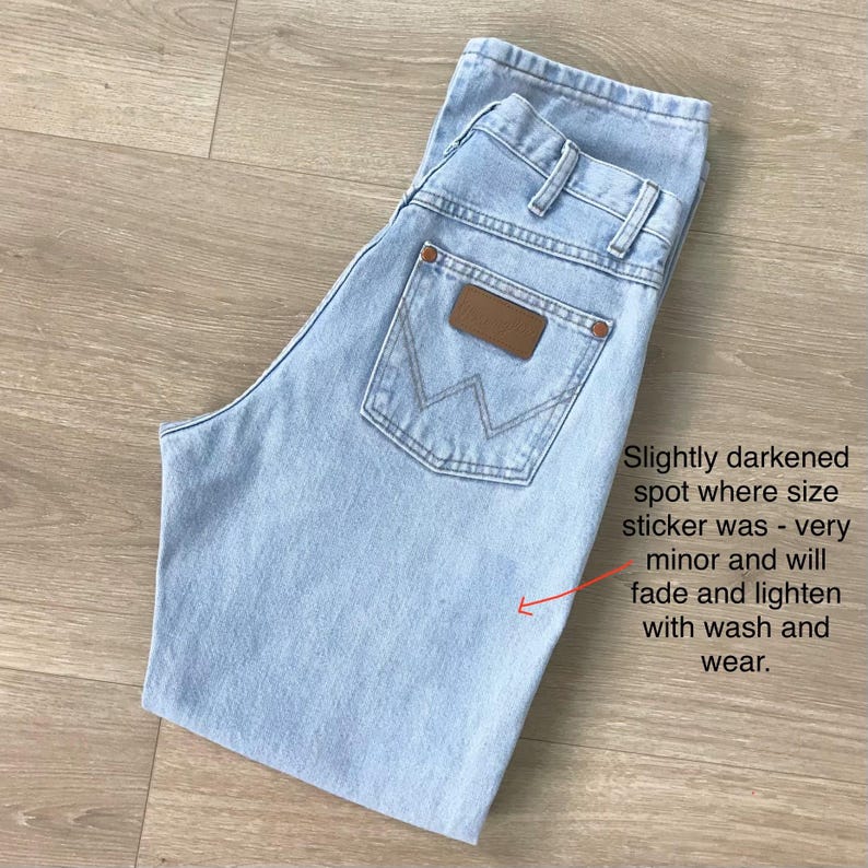 May include: Pale blue denim jeans with a brown leather patch on the back pocket. The jeans have a classic five-pocket design and are folded. Text on the image: "Slightly darkened spot where size sticker was - very minor and will fade and lighten with wash and wear."