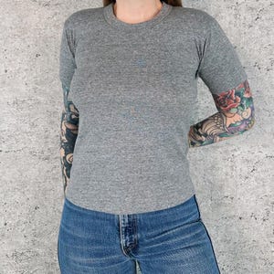 May include: A heather gray crew neck t-shirt. The short-sleeved shirt is made of a soft material. The model is wearing blue jeans. The background is a light gray textured wall.