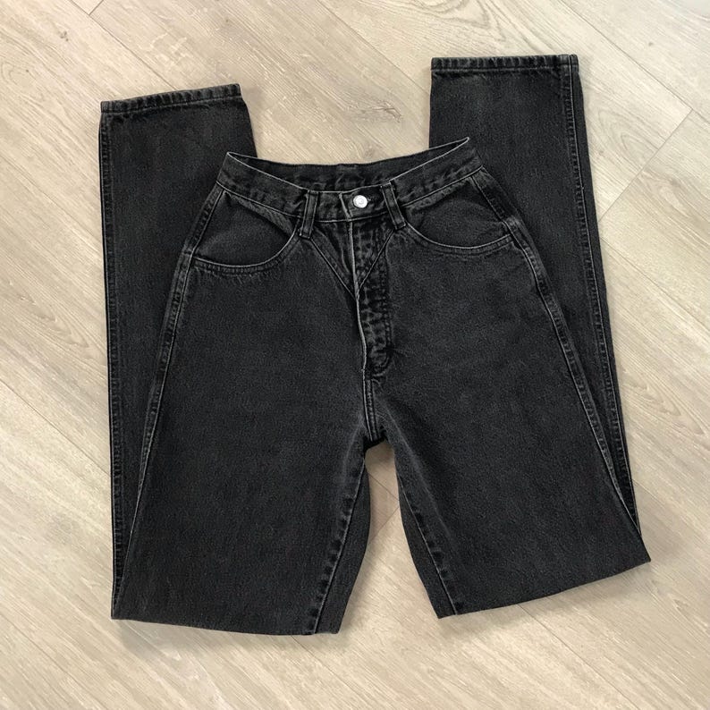 Pu&ograve; includere: Un paio di jeans neri in denim, stesi piatti. I jeans hanno una vita alta, chiusura con bottoni e un classico design a cinque tasche. Il tessuto sembra essere un denim di peso medio, con dettagli di cucitura visibili. I jeans sono di colore nero pieno.