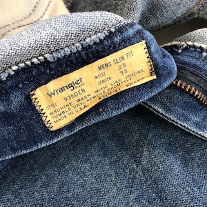 May include: Close-up of a pair of blue denim Wrangler jeans. The label reads "MENS SLIM FIT" with waist size 29 inches and length 33 inches. The label also includes washing instructions and "MADE IN U.S.A."