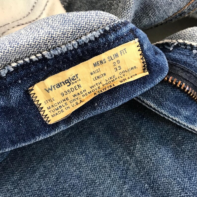 May include: Close-up of a pair of blue denim Wrangler jeans. The label reads "MENS SLIM FIT" with waist size 29 inches and length 33 inches. The label also includes washing instructions and "MADE IN U.S.A."