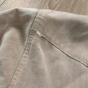May include: Close-up of a tan-colored garment, showing the shoulder and collar area. The fabric has a textured appearance with visible stitching along the seams. A small area of fraying is visible near the collar.