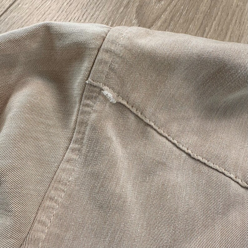 May include: Close-up of a tan-colored garment, showing the shoulder and collar area. The fabric has a textured appearance with visible stitching along the seams. A small area of fraying is visible near the collar.