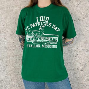 Op de afbeelding: Groen t-shirt met witte tekst: "I DID ST. PATRICK'S DAY AT R.U. GRUMPY'S Beer-Booze-Bands & Big Times O'FALLON, MISSOURI". Het shirt heeft een ronde hals.