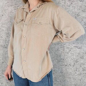 May include: A tan button-down shirt with two chest pockets and snap closures. The shirt has long sleeves and a collared neckline. A small label is visible on the chest pocket. The shirt is paired with blue jeans.