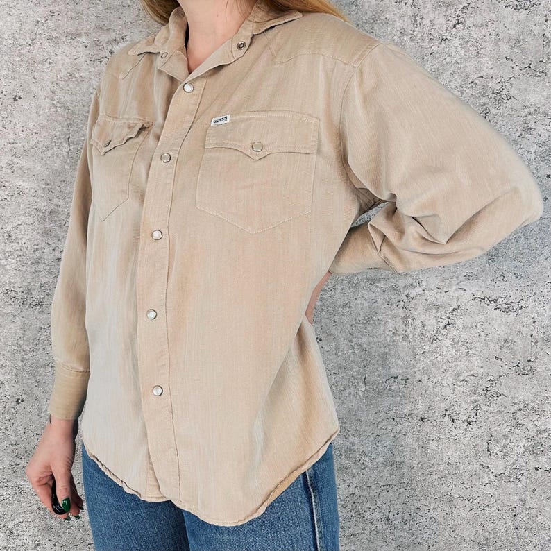 May include: A tan button-down shirt with two chest pockets and snap closures. The shirt has long sleeves and a collared neckline. A small label is visible on the chest pocket. The shirt is paired with blue jeans.