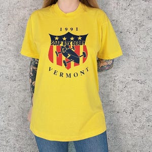 May include: Yellow t-shirt with a graphic featuring the text "1991 SOAP BOX DERBY VERMONT". The graphic includes a shield design with stars, red stripes, and a soap box derby car.