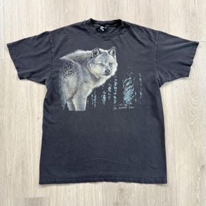 Peut inclure: T-shirt noir avec une illustration d&eacute;taill&eacute;e d'un loup gris et d'une for&ecirc;t. Le texte "St. Louis Zoo" est imprim&eacute; sous les arbres. Le t-shirt est fait d'une mati&egrave;re douce.