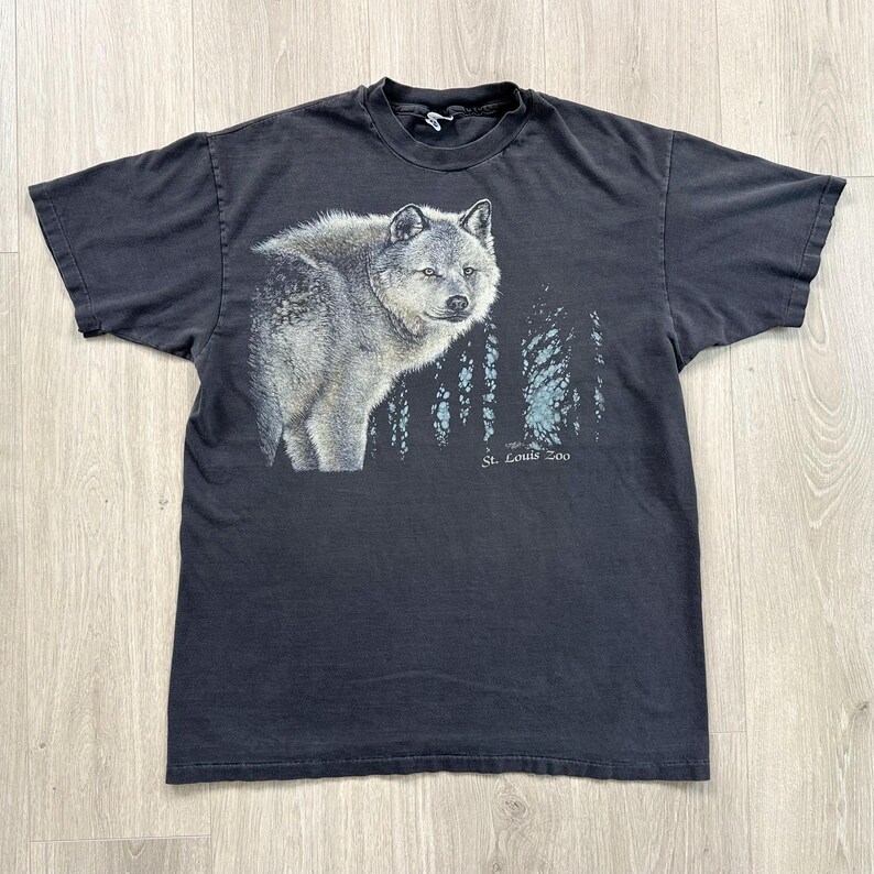 Peut inclure: T-shirt noir avec une illustration d&eacute;taill&eacute;e d'un loup gris et d'une for&ecirc;t. Le texte "St. Louis Zoo" est imprim&eacute; sous les arbres. Le t-shirt est fait d'une mati&egrave;re douce.