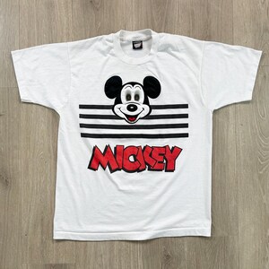 May include: White t-shirt featuring a graphic of Mickey Mouse with black ears, a smiling face, and the name "Mickey" in red block letters. The design includes black horizontal stripes.