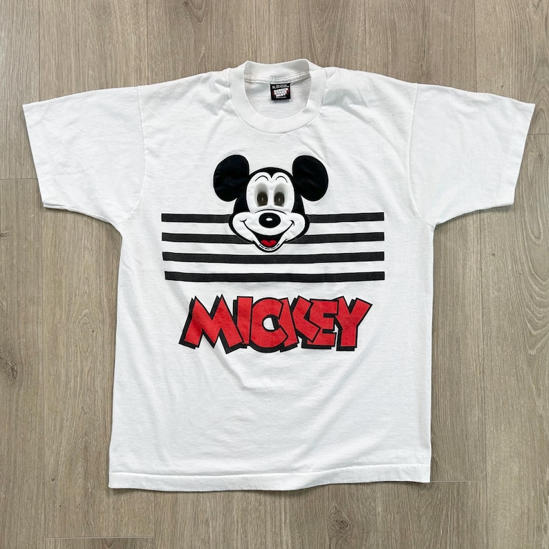 May include: White t-shirt featuring a graphic of Mickey Mouse with black ears, a smiling face, and the name "Mickey" in red block letters. The design includes black horizontal stripes.