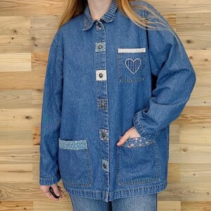 May include: A blue denim jacket with a collared neckline and button-up closure. The jacket features decorative patches, including a white embroidered heart on the pocket. The pockets and collar have patterned fabric accents.