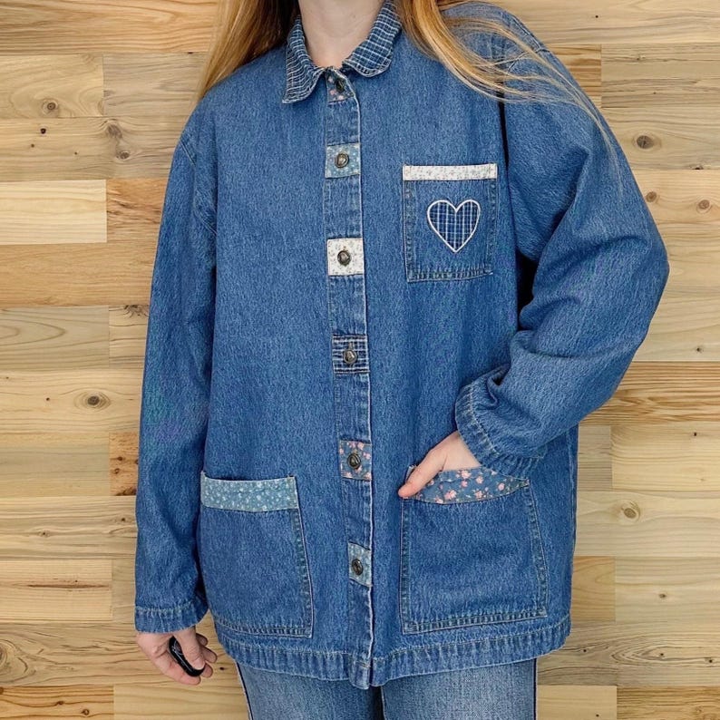 May include: A blue denim jacket with a collared neckline and button-up closure. The jacket features decorative patches, including a white embroidered heart on the pocket. The pockets and collar have patterned fabric accents.