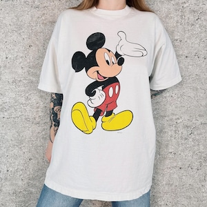 Op de afbeelding: Wit T-shirt met een grote Mickey Mouse-afbeelding. Het stripfiguur draagt een rode broek, gele schoenen en witte handschoenen. Mickey lacht en zwaait met zijn rechterhand. Het T-shirt heeft korte mouwen.