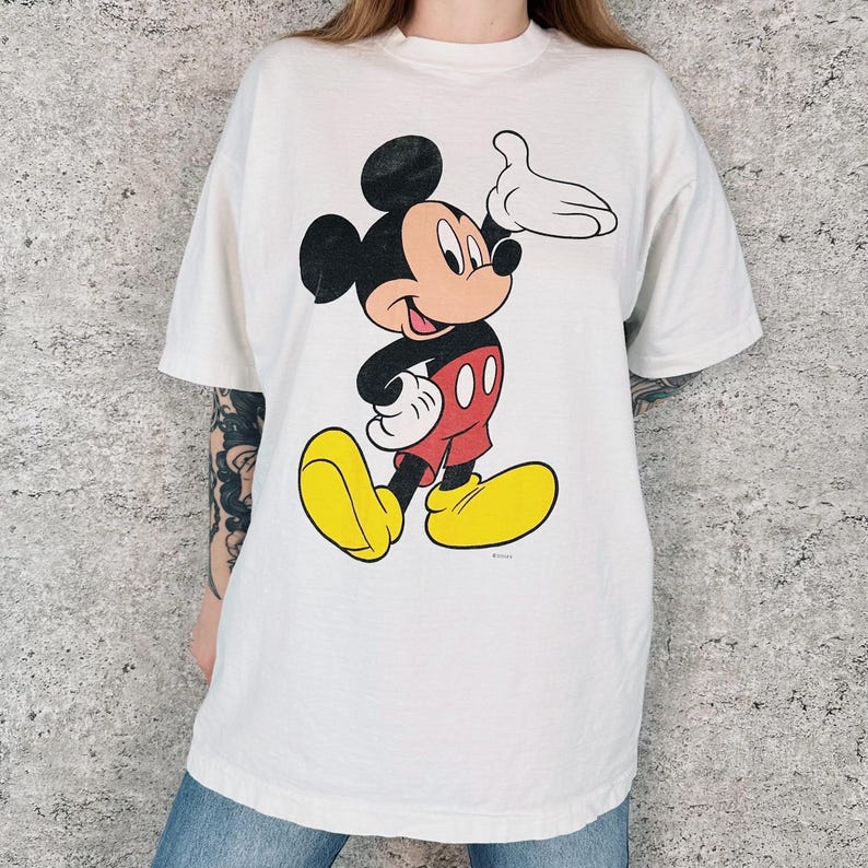 Op de afbeelding: Wit T-shirt met een grote Mickey Mouse-afbeelding. Het stripfiguur draagt een rode broek, gele schoenen en witte handschoenen. Mickey lacht en zwaait met zijn rechterhand. Het T-shirt heeft korte mouwen.