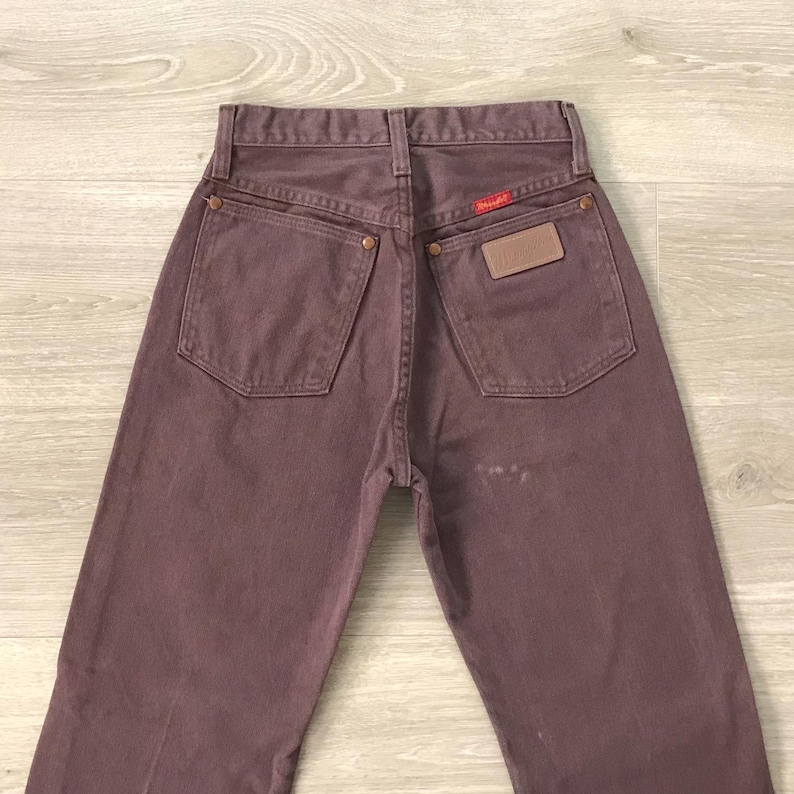 Op de afbeelding: Een paar vintage Wrangler jeans in mauve. De jeans hebben een klassiek ontwerp met vijf zakken, een rood Wrangler-label en een bruine leren patch op de rug. De jeans zijn gemaakt van duurzaam denim.