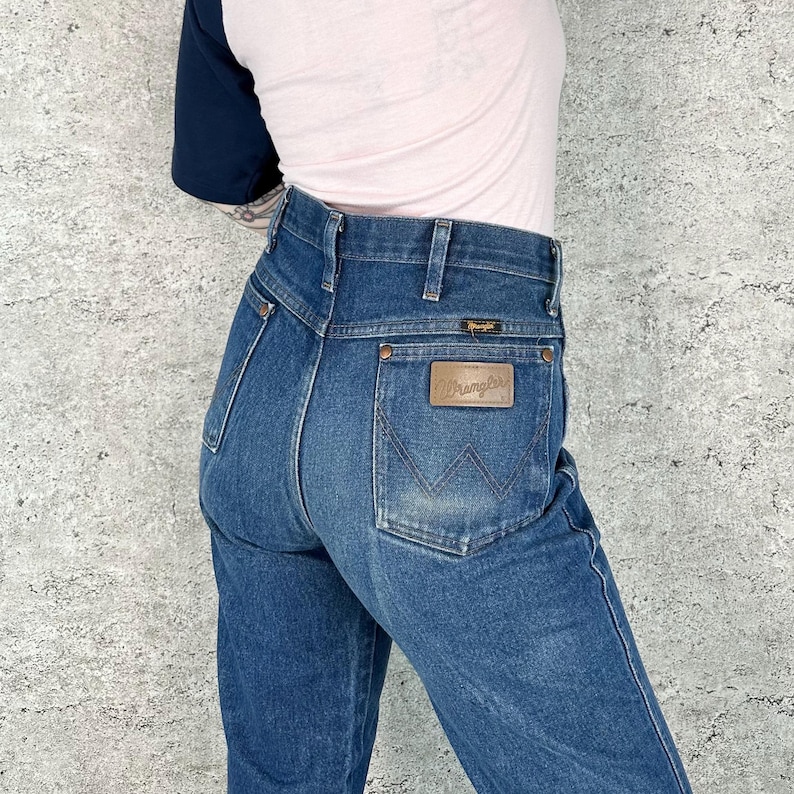 May include: Dark wash denim Wrangler jeans. The jeans have a high waist and a classic five-pocket design. A brown leather patch with the Wrangler logo is on the back pocket. The jeans are worn with a light pink and navy blue top.