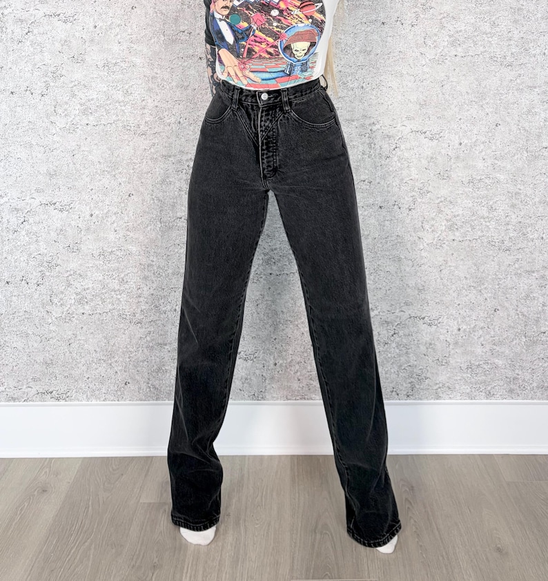 Pu&ograve; includere: Jeans neri a vita alta con gamba dritta. I jeans hanno una chiusura con bottoni e cerniera e un classico design a cinque tasche. Il denim sembra essere un lavaggio scuro e i jeans sono indossati con una maglietta grafica.