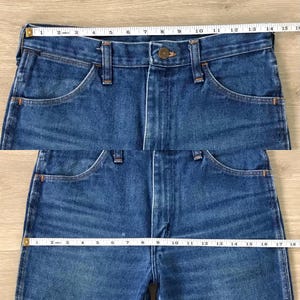 May include: Close-up of blue denim jeans with a button closure and pockets. A measuring tape shows the waist size. The jeans have a classic design and are made of durable denim fabric.