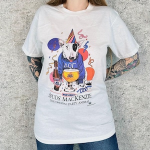 May include: White t-shirt featuring a cartoon Bull Terrier wearing a blue sweater with Greek letters, a party hat, and holding a beer glass. The shirt has the text "Spuds MacKenzie The Original Party Animal."