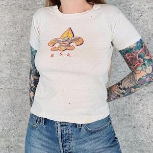 May include: Cream-colored t-shirt featuring a colorful BSA emblem with the text "B. S. A." in red. The shirt has a crew neck and short sleeves. The shirt is paired with blue denim jeans.