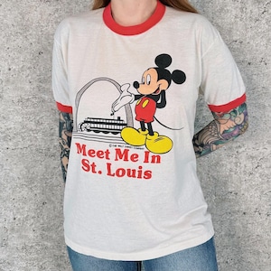 May include: White t-shirt with red trim featuring a graphic of Mickey Mouse in front of a St. Louis landmark. The text "Meet Me In St. Louis" is printed in red. The shirt has short sleeves and a crew neck.