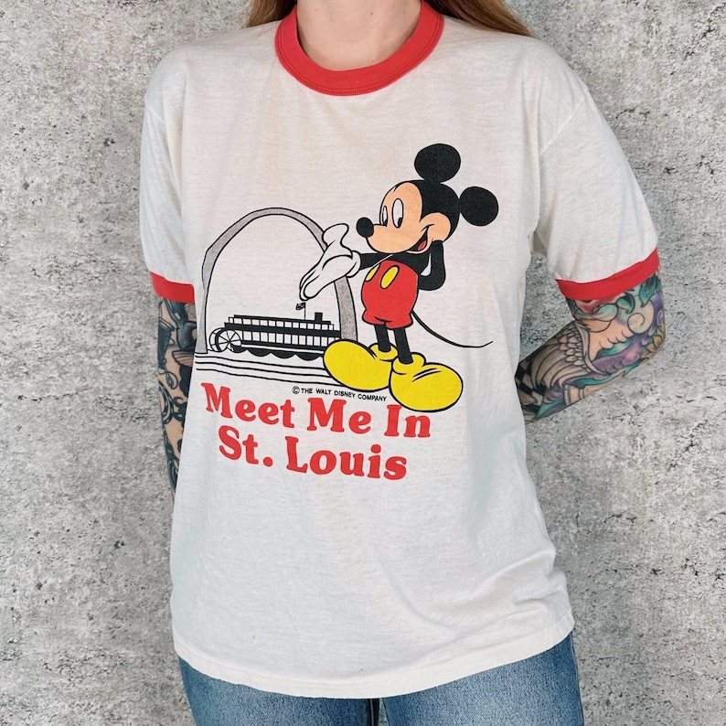 May include: White t-shirt with red trim featuring a graphic of Mickey Mouse in front of a St. Louis landmark. The text "Meet Me In St. Louis" is printed in red. The shirt has short sleeves and a crew neck.