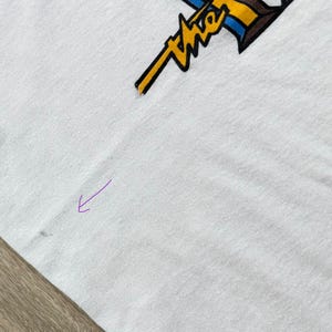 May include: White t-shirt with a graphic design in yellow, brown, and blue. The design features the word "The" in a stylized font. The shirt is made of a soft, likely cotton material.